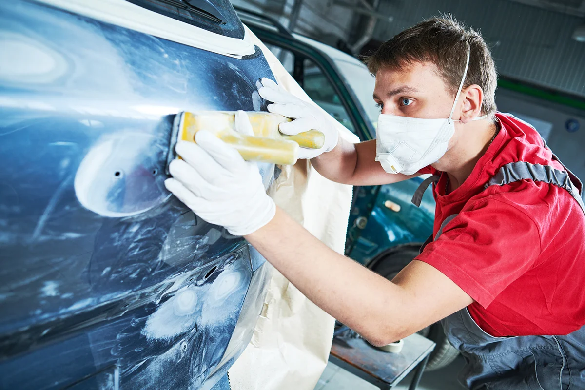 Auto body technician sanding a car panel while wearing a protective mask and gloves in a repair shop.